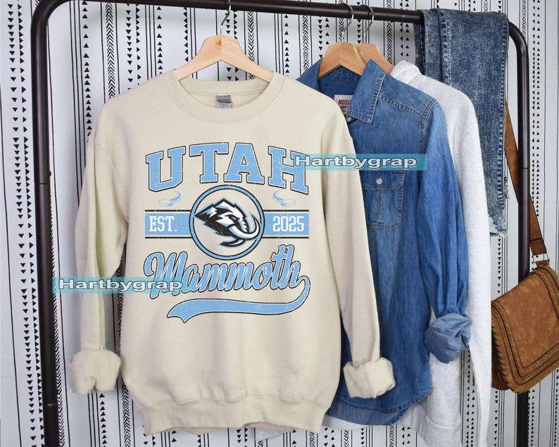 May include: Cream-colored sweatshirt with the text "UTAH Mammoth EST. 2025" in light blue. The design includes a circular emblem with a mammoth graphic. Other clothing items are visible in the background.