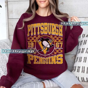 Pu&ograve; includere: Felpa bordeaux con la scritta "PITTSBURGH PENGUINS" in lettere dorate e un logo della squadra di hockey. Il logo include un pinguino con una mazza da hockey e i numeri "19" e "67".