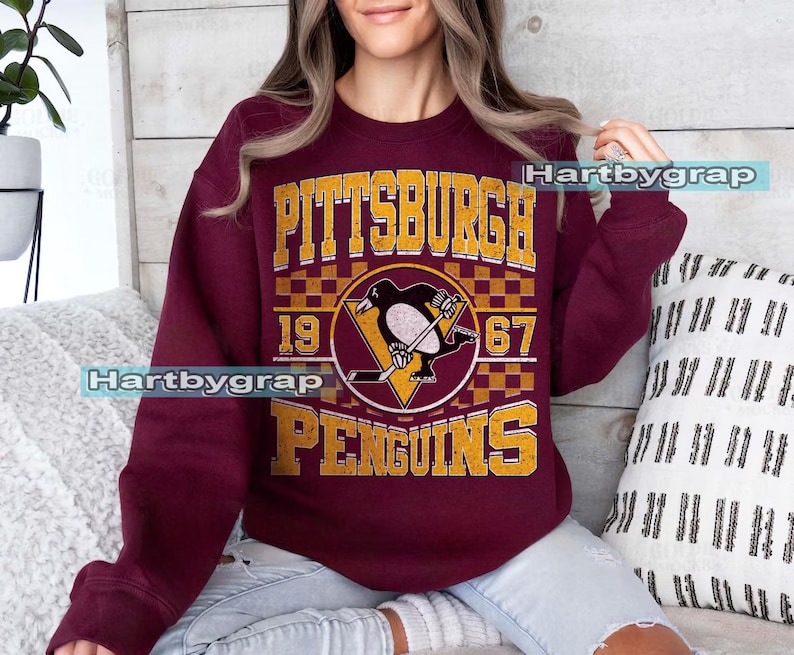 Pu&ograve; includere: Felpa bordeaux con la scritta "PITTSBURGH PENGUINS" in lettere dorate e un logo della squadra di hockey. Il logo include un pinguino con una mazza da hockey e i numeri "19" e "67".