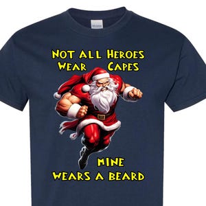 Puede incluir: Camiseta azul marino con un diseño de un Papá Noel musculoso con traje y gorro rojos. El texto dice "NOT ALL HEROES WEAR CAPES, MINE WEARS A BEARD" en amarillo. Un diseño divertido y festivo.