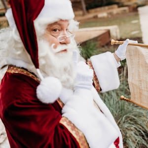 Pode incluir: Pai Natal em fato e chapéu de veludo vermelho, segurando um pergaminho com uma lista. Ele usa luvas brancas e óculos, com barba e bigode longos e brancos. O pergaminho é bege com texto.