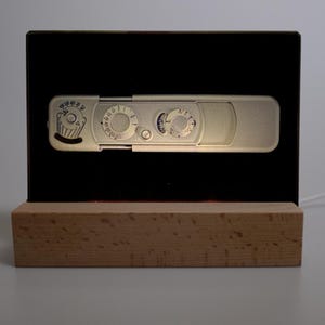 May include: A silver-colored camera-shaped light fixture with dials and a cupcake graphic is mounted on a black background. The light sits on a rectangular wooden base. A white cord extends from the base.