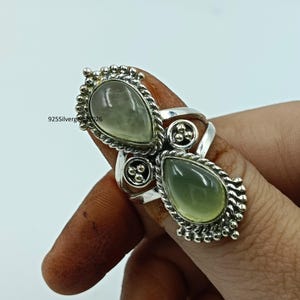 May include: A silver ring featuring two pear-shaped, pale green gemstones. The ring has a detailed, ornate design with a beaded border around the stones and decorative accents. The text "925Silvergems026" is visible.