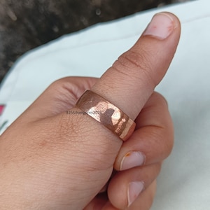 Pode incluir: Um anel cor de cobre com uma superfície texturizada é usado em um polegar. O anel tem um design em forma de faixa e uma textura ligeiramente irregular. O texto "925silvergems2026" é visível no anel.