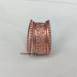Op de afbeelding: Een koperkleurige ring met een textuurontwerp. De ring heeft een centrale band met een geëtst geometrisch patroon, geflankeerd door gedraaide touwachtige banden. De tekst "925Silvergems2026" is zichtbaar.