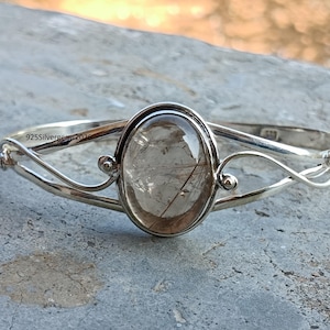 Peut inclure: Un bracelet jonc en argent avec une pierre précieuse ovale transparente et des inclusions sombres. Le bracelet a un design délicat et ouvert avec des lignes courbes et de petits accents décoratifs. Le texte "925silvergems2020" est visible.