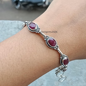 May include: A silver bracelet with oval, deep red gemstones. The bracelet features an ornate design with the gemstones set in decorative bezels. The bracelet is on a wrist, with the text "Silvergems2026" visible.