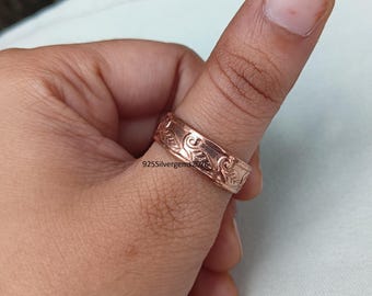 Könnte beinhalten: Ein roségoldener Ring mit einem aufwendigen, sich wiederholenden Blumenmuster. Der Ring wird an einem Finger getragen, wodurch das detaillierte Design zur Geltung kommt. Der Ring ist wahrscheinlich aus einem Edelmetall gefertigt und ein Schmuckstück.
