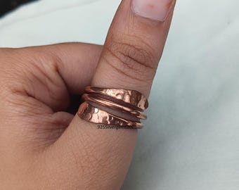 Op de afbeelding: Een koperkleurige ring met een gehamerde textuur is om een duim gewikkeld. De ring heeft meerdere banden en een spiraalvormig ontwerp. De tekst "925Silvergems2026" is zichtbaar op de ring.