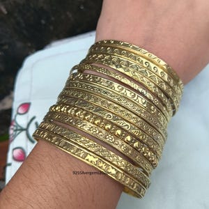 Puede incluir: Una pila de pulseras doradas con intrincados diseños grabados. Las pulseras están apiladas en una muñeca, mostrando varios patrones, incluyendo motivos florales y geométricos. La estética general es ornamentada y decorativa.
