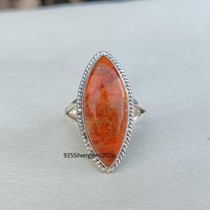 May include: A silver ring featuring an oval, orange gemstone. The gemstone is set in a silver band with a twisted rope design around the stone. The ring has a simple band and is displayed on a white surface. The text "925Silvergems2026" is visible.