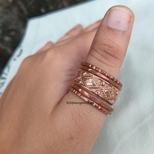 Könnte beinhalten: Kupferfarbenes Ring-Set auf einem Daumen. Die Ringe haben ein strukturiertes Design mit geometrischen und floralen Mustern. Die Ringe sind übereinander gestapelt und bilden ein einzigartiges Accessoire. Die Ringe haben eine warme, rötlich-braune Farbe.