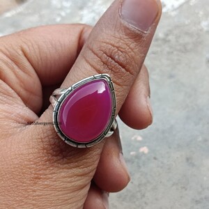 May include: A silver ring with a large, teardrop-shaped, magenta gemstone. The gemstone is set in a silver bezel with a textured border. The ring is on a person's finger.
