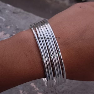 Op de afbeelding: Een stapel glanzende zilveren armbanden om een pols. De armbanden zijn dun en rond, en reflecteren het licht. De afbeelding bevat de tekst "925silvergems2026".