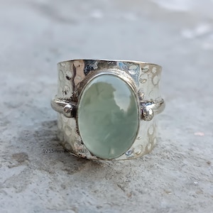 Puede incluir: Un anillo ancho de plata con una banda texturizada y una gema ovalada de color verde pálido. El anillo tiene un diseño de metal martillado y el texto "925Silvergems2026" es visible en la banda.