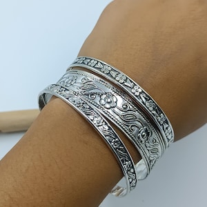 Puede incluir: Un conjunto de brazaletes de plata con diseños florales y de hojas. Los brazaletes están apilados en una muñeca, mostrando los intrincados detalles de los diseños grabados. El metal tiene una superficie pulida y reflectante.