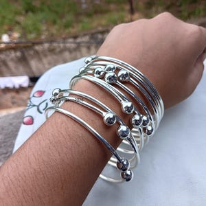 May include: A collection of silver bangles with a simple design. Each bangle features a smooth, rounded ball accent. The bangles are stacked on a wrist, showcasing their reflective, polished surface. The jewelry is displayed against a soft, neutral background.