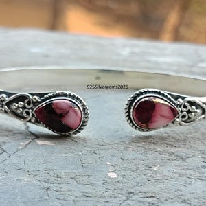 Può includere: Un bracciale a polsino in argento con due pietre a forma di goccia, rosa e marrone. Il bracciale presenta dettagli decorativi attorno alle pietre e il testo "925silvergems2026".