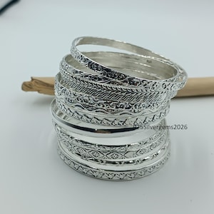May include: A stack of silver bangles with intricate patterns. The bangles have various designs, including floral and geometric motifs. The jewelry is displayed on a white surface with a wooden stick in the background. The text "925Silvergems2026" is visible.