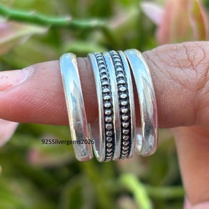 May include: A collection of silver rings, including smooth bands and a beaded design, are stacked on a finger. The rings are displayed against a blurred green and pink background. The text "925Silvergems2026" is visible.