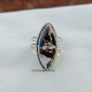 Può includere: Un anello d'argento con una pietra di forma ovale. La pietra presenta un mix di colori, tra cui nero, bianco, blu e rame. L'anello ha un bordo intrecciato attorno alla pietra e un design a tripla fascia. Il testo "925Silvergems2026" è visibile.