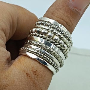Puede incluir: Una pila de anillos de plata de varios diseños, incluyendo bandas con cuentas, trenzadas y estampadas. Los anillos se llevan en un dedo, mostrando las diferentes texturas y estilos de la joyería.