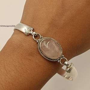 Könnte beinhalten: Ein silbernes Armband mit einem ovalen Rosenquarz-Edelstein. Der Edelstein ist in einer silbernen Lünette mit einem gedrehten Seilmuster gefasst. Das Armband hat ein Scharnierdesign und wird an einem Handgelenk gezeigt.