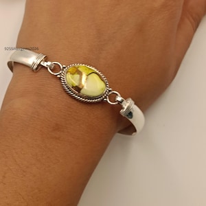 Puede incluir: Una pulsera de plata con una piedra ovalada multicolor. La piedra tiene tonos de amarillo, verde y marrón. La pulsera está en una muñeca. El texto "925Silvergems2026" es visible.