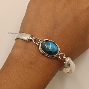 May include: A silver bracelet featuring an oval labradorite gemstone with blue and green iridescence. The gemstone is set in a decorative bezel with a twisted rope design. The bracelet has a hinged design and is connected with small silver links.