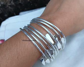 Puede incluir: Un conjunto de brazaletes plateados con un diseño único. Los brazaletes se llevan en la muñeca y presentan una serie de bandas curvas y abiertas con formas geométricas. El texto "925Silvergems2026" es visible.