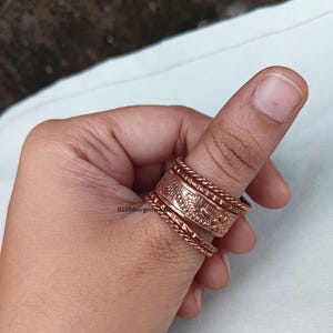 Könnte beinhalten: Ein Satz kupferfarbener Ringe an einem Daumen. Die Ringe haben verschiedene Designs, darunter florale Muster und gedrehte Seiltexturen. Die Ringe sind übereinander gestapelt, wodurch ein geschichteter Effekt entsteht. Der Text "925silvergems" ist sichtbar.