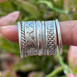 Puede incluir: Un conjunto de anillos de plata con diversos diseños, incluyendo cuerdas trenzadas, motivos florales y bandas texturizadas. Los anillos se muestran en un dedo, mostrando sus intrincados detalles y brillo. El texto "925Silvergems2026" es visible.