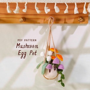 May include: A crocheted mushroom egg pot hanging from a wooden rack. The pot is filled with colorful crocheted mushrooms in white, orange, and purple. The text "PDF PATTERN Mushroom Egg Pot" is visible.