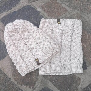 Può includere: Un set coordinato di cappello e scaldacollo in maglia beige chiaro. Entrambi gli articoli presentano un motivo strutturato e intrecciato, impreziositi da piccole etichette in pelle marrone. Il set è progettato per calore e stile.