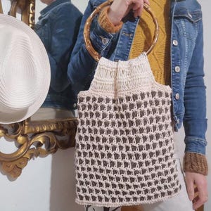 Può includere: Borsa tote all'uncinetto fatta a mano con manico in bambù. La borsa presenta una parte superiore color crema e un corpo a fantasia marrone e crema. Il manico è marrone chiaro. La borsa è tenuta da una persona con una giacca di jeans.
