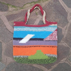 Può includere: Una colorata borsa tote all'uncinetto fatta a mano. La borsa presenta strisce orizzontali in vari colori, tra cui arancione, blu, viola e verde. Il design include forme geometriche bianche e rosse. I manici sono in tessuto rosso e oro ritorto.