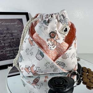 May include: A drawstring bag with a white background and a pattern of foxes, owls, and trees in gray and peach. The bag is open, revealing a peach-colored interior. A framed print of chamomile benefits is in the background.