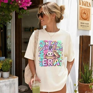 May include: A cream-colored short-sleeve t-shirt featuring a colorful graphic print of a cute dumpling in a bamboo steamer with the text "IN MY DUMPLING ERA" surrounded by small stars, hearts, and swirls in shades of pink, blue, and yellow.