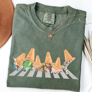 May include: Olive green t-shirt featuring a graphic of cartoon characters crossing a crosswalk under orange traffic cones. The shirt has a Comfort Colors tag and a silver necklace with a small pendant. A brown hat is visible in the background.