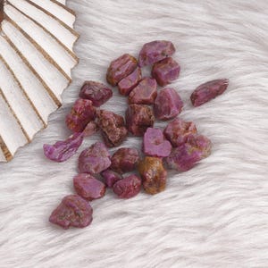 Op de afbeelding: Een verzameling ruwe, ongeslepen robijn edelstenen in verschillende maten en vormen. De edelstenen vertonen een dieprode tot paarse tint met natuurlijke texturen. Ze liggen op een zacht, wit imitatiebont oppervlak, met een decoratieve houten waaier op de achtergrond.