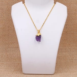 Pode incluir: Um colar de corrente dourada com um pingente de ametista roxa bruta. A ametista tem uma tampa dourada. O colar é exibido em um busto branco sobre um fundo neutro.