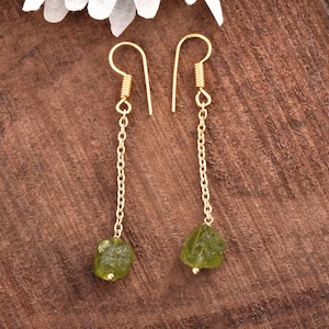 May include: A pair of gold-colored dangle earrings. Each earring features a gold-colored chain with a rough, green gemstone at the bottom. The earrings are displayed on a brown wooden surface.