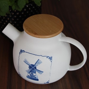 Op de afbeelding: Witte keramische theepot met een houten deksel. De theepot heeft een blauw windmolenontwerp in een decoratief kader. De handgreep en de tuit zijn wit. Een zwart-wit patroonpot met groene bladeren staat op de achtergrond.