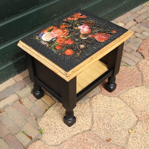 Op de afbeelding: Kleine, vierkante bijzettafel met zwart frame en poten. Het tafelblad heeft een bloemenontwerp met rode, oranje en roze bloemen op een zwarte achtergrond. De tafel heeft een onderste plank en een natuurlijke houten rand.