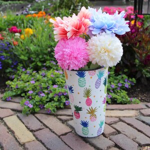 Könnte beinhalten: Eine weiße Vase mit Ananasmuster enthält einen Strauß bunter Kunstblumen. Die Blumen sind rosa, korallenfarben, blau und weiß. Die Vase steht auf einem Ziegelweg, mit einem Garten im Hintergrund.