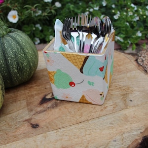 May include: A square utensil holder with an ice cream cone design, filled with cutlery. Two green gourds are on a wooden surface beside the holder. The background shows greenery and flowers.