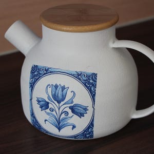 May include: White ceramic teapot with a wooden lid and a curved handle. The teapot features a blue floral design on the front. The spout is angled, and the design is reminiscent of Delftware.