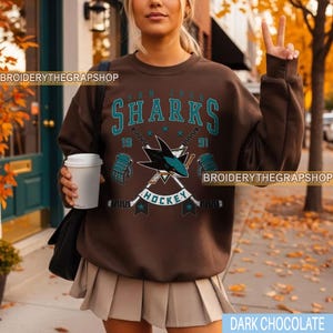 Puede incluir: Una sudadera de cuello redondo color marr&oacute;n oscuro con un gr&aacute;fico en azul cerceta y blanco del logotipo de los San Jose Sharks, palos de hockey cruzados y el texto San Jose Sharks Hockey 1991, incluyendo dos guantes y estrellas.