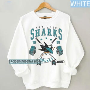 Puede incluir: Una sudadera blanca de cuello redondo con un gr&aacute;fico desgastado del logotipo de los San Jose Sharks, palos de hockey cruzados y guantes. El texto dice San Jose Sharks 1991 Hockey. El dise&ntilde;o utiliza colores verde azulado, negro y blanco.