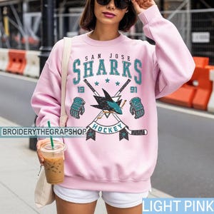 Puede incluir: Una sudadera de cuello redondo rosa claro con un estampado gr&aacute;fico del logotipo de los San Jose Sharks con palos de hockey cruzados, guantes y el texto San Jose Sharks 1991 Hockey. El dise&ntilde;o utiliza colores verde azulado, negro y blanco.
